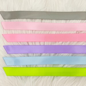 Pode incluir: Uma cole&ccedil;&atilde;o de fitas de cetim coloridas em v&aacute;rios tons. As fitas incluem cinza, rosa claro, rosa m&eacute;dio, lavanda, azul claro e verde neon. As fitas est&atilde;o dispostas horizontalmente sobre um fundo branco e fofo.