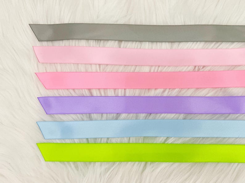 Pode incluir: Uma cole&ccedil;&atilde;o de fitas de cetim coloridas em v&aacute;rios tons. As fitas incluem cinza, rosa claro, rosa m&eacute;dio, lavanda, azul claro e verde neon. As fitas est&atilde;o dispostas horizontalmente sobre um fundo branco e fofo.