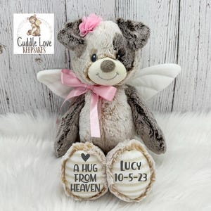 May include: A grey and white plush dog with pink accents and angel wings. The dog has a pink bow around its neck and the words "A hug from heaven" and "Lucy 10-5-23" on its paws.