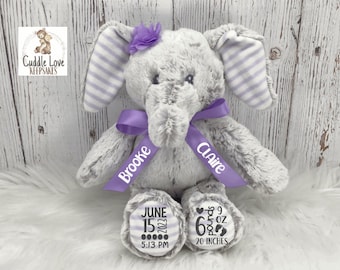 May include: Gray plush elephant stuffed animal with purple striped ears and a purple bow. The elephant has personalized embroidered names "Brooke" and "Claire" on the bow. The elephant's feet have embroidered details including the date "June 15, 2022", time "5:13 PM", weight "6 lbs 9 oz", and length "20 inches".
