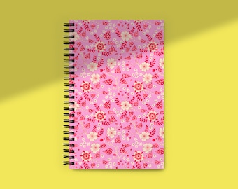 May include: A spiral-bound notebook with a pink cover featuring a floral pattern in red, white, and cream. The notebook is set against a yellow background, with a shadow cast across the surface.