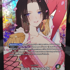 May include: A holographic One Piece trading card featuring Boa Hancock with long black hair, blue eyes, and snake-shaped earrings. The card displays the text 'Leader Boa Hancock', '5000' power, and game rules against a background of pink cherry blossoms.
