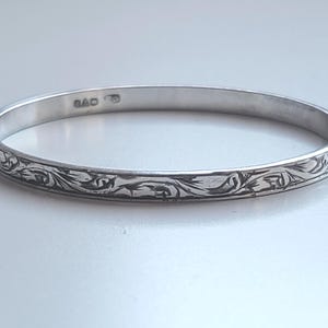 May include: A silver bangle bracelet with an engraved floral pattern. The bracelet is round and has a polished finish. The engraved design wraps around the entire bracelet. The bracelet is approximately 0.6 cm wide.