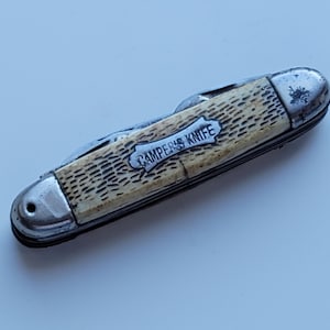 Könnte beinhalten: Ein klappbares Taschenmesser mit einem strukturierten, cremefarbenen Griff und einer silberfarbenen Metalleinlage, in die der Text CAMPER'S KNIFE eingraviert ist. Das Messer hat polierte silberne Metallbacken an beiden Enden und eine Klinge.