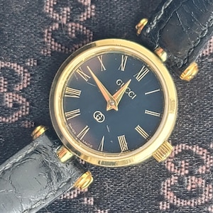 May include: A gold-tone wristwatch with a round case and a black dial featuring gold Roman numeral hour markers. The watch has gold hands, the Gucci logo at the top, a small GG emblem above the six o'clock position, and a black leather strap.