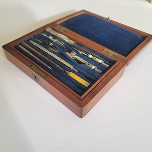 Op de afbeelding: Een houten set tekeninstrumenten in een rechthoekige doos met een scharnierend deksel. De binnenkant is bekleed met blauw fluweel en bevat diverse metalen passers, een verdeelpasser en een geel potlood in op maat gemaakte uitsparingen.