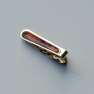 May include: A gold-toned metal tie clip featuring a rectangular inlay of deep red, translucent material. The tie clip has a polished, reflective finish and a spring-loaded mechanism on the underside for securing it to a necktie.