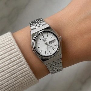 May include: A silver Seiko automatic wristwatch with a white face and a silver strap. The watch face displays the day and date. The watch is on a wrist with a beige sweater sleeve.
