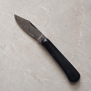 Könnte beinhalten: Ein klappbares Taschenmesser mit einer einzelnen, verwitterten Metallklinge, die sichtbare Markierungen nahe der Klinge aufweist, und einem glatten, einfarbig schwarzen Griff. Die Klinge zeigt Gebrauchsspuren mit einer Spitze.