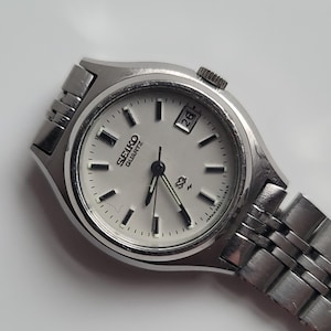 May include: A silver-toned Seiko Quartz wristwatch featuring a round white dial with black hour markers, silver hands, and a date window at the three o'clock position. The watch includes a metallic link bracelet and a crown on the right side.