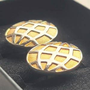 Puede incluir: Un par de gemelos ovalados. Los gemelos tienen un interior dorado con un diseño de rejilla plateada superpuesto. Los gemelos se presentan en una caja negra, lo que sugiere que son una joya.