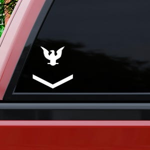 May include: White vinyl decal of a military rank insignia, featuring an eagle above a chevron. The decal is affixed to the rear window of a red pickup truck. The background includes green foliage.