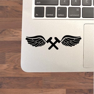 May include: Black and white sticker of crossed hammers with wings. The sticker is on a silver laptop.