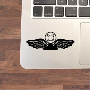 May include: Black vinyl sticker featuring a stylized emblem with wings and a geometric center design. The sticker is placed on a laptop, with keyboard keys visible in the background. The sticker's design is a bold, graphic element.