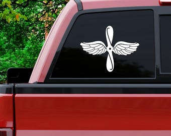 May include: A white vinyl decal of a propeller with wings, commonly used as a military aviation symbol. The decal is applied to the rear window of a red pickup truck.