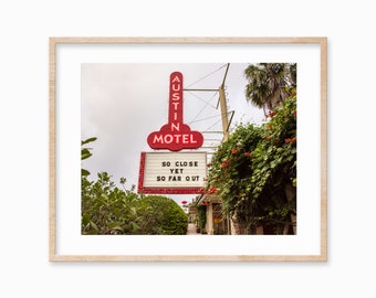 May include: A vintage motel sign with red lettering that reads "Austin Motel" and a message board that says "So Close Yet So Far Out". The sign is surrounded by lush green foliage.