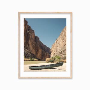 May include: Framed print of a scenic landscape featuring a green canoe resting on a sandy riverbank. Towering rock formations flank the river under a clear blue sky. The artwork is encased in a light-colored wooden frame.