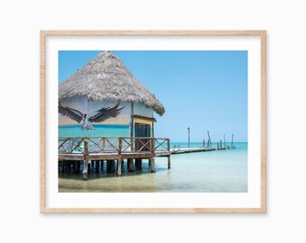 May include: A small, thatched-roof hut on stilts over turquoise water. The hut has a mural of a bird in flight on the side. A wooden pier extends from the hut into the water.
