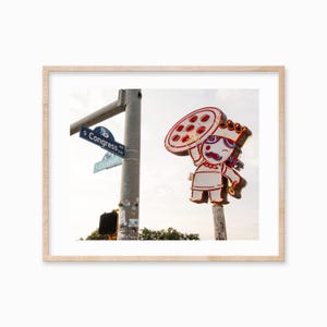 May include: A neon sign featuring a cartoon character holding a pizza. The sign is mounted on a pole and is situated at a street junction. The street signs read "S Congress Ave" and "E Elizabeth St."