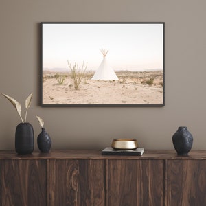 Puede incluir: Una fotografía enmarcada de una tienda de campaña tipi blanca en un paisaje desértico. El tipi está rodeado de plantas del desierto y el cielo es de un azul claro. La foto está colgada en una pared sobre un armario de madera con un cuenco dorado y jarrones negros.