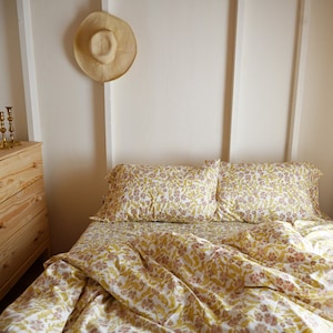 May include: A white bed with a yellow and pink floral patterned duvet cover and matching pillowcases. The bed is made and the duvet is slightly rumpled. A straw hat hangs on the wall behind the bed.