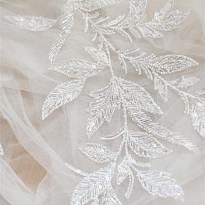 Könnte beinhalten: Weiße Perlen-Spitzenborte mit einem zarten Blumenmuster. Die Borte ist mit kleinen weißen Perlen und Pailletten verziert, die in einem Muster aus Blättern und Zweigen angeordnet sind.