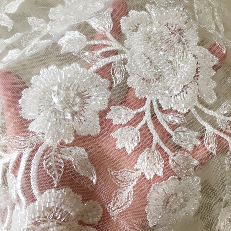 May include: White beaded floral lace fabric with a delicate mesh background. The fabric features intricate floral designs with small, white beads.