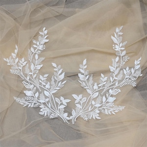 May include: Two white floral lace appliques. Each applique features a delicate design of leaves and small beaded accents. The appliques are arranged on a sheer, beige fabric, creating a soft, elegant look. Ideal for bridal or craft projects.