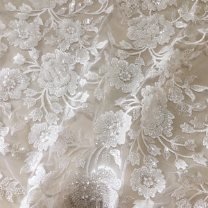 May include: White floral lace fabric with beaded embellishments. The lace features a delicate floral pattern with small, white beads scattered throughout.