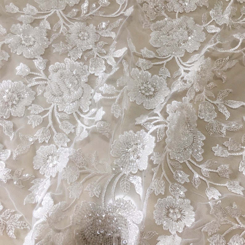 May include: White floral lace fabric with beaded embellishments. The lace features a delicate floral pattern with small, white beads scattered throughout.