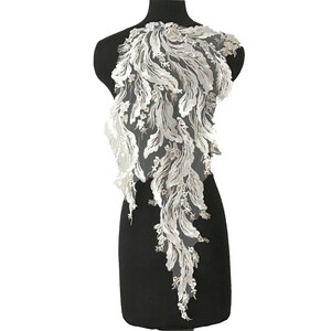 Może przedstawiać: Biała aplikacja z koronki z delikatnym wzorem kwiatowym. Koronka jest ułożona w kaskadowym wzorze, przypominającym pióra lub wodospad.