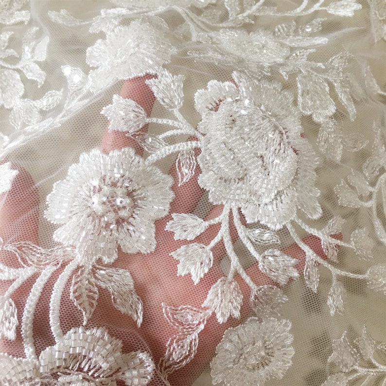 May include: White floral lace fabric with delicate beading and a sheer mesh background. The lace features a repeating pattern of flowers and leaves.