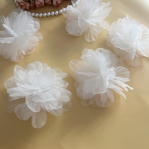 Puede incluir: Seis flores blancas de tela con un aspecto delicado y aireado. Los pétalos son suaves y translúcidos, creando un aspecto ligero y etéreo. Una hilera de perlas y pequeñas flores rosas están en el fondo. Adecuadas para manualidades y decoraciones.