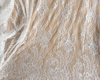 May include: White lace fabric with a floral pattern. The delicate lace is draped, revealing the texture and intricate details of the design. The fabric is suitable for bridal wear, dresses, or other craft projects.
