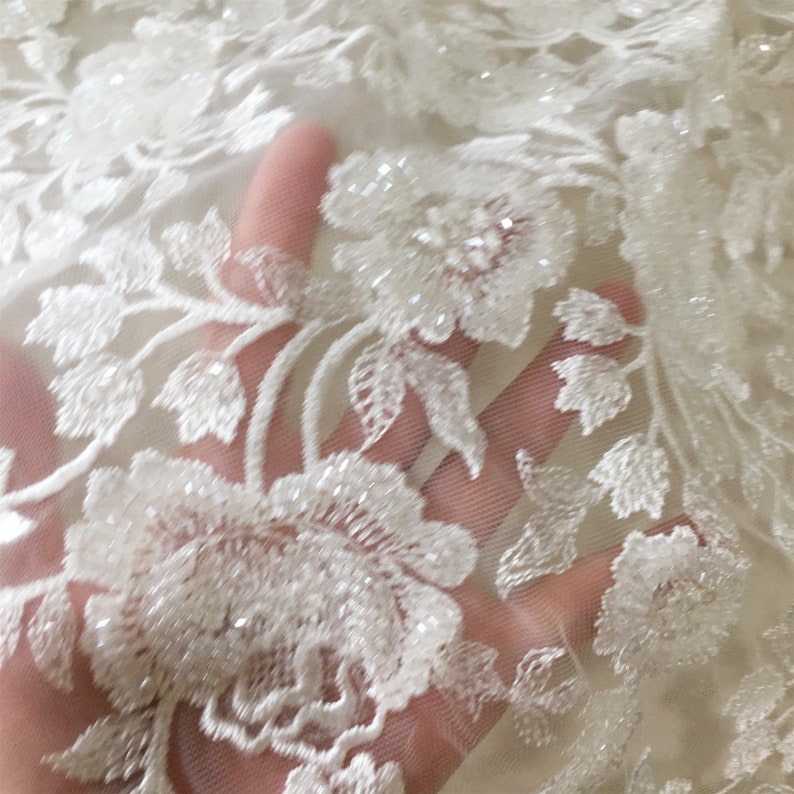May include: White beaded floral lace fabric with a delicate pattern. The lace is sheer and has a soft, flowing texture.