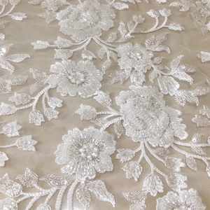 May include: White beaded floral lace fabric with a delicate pattern. The lace is made of small white beads that are sewn together to create a floral design. The fabric is sheer and has a soft, flowing texture.