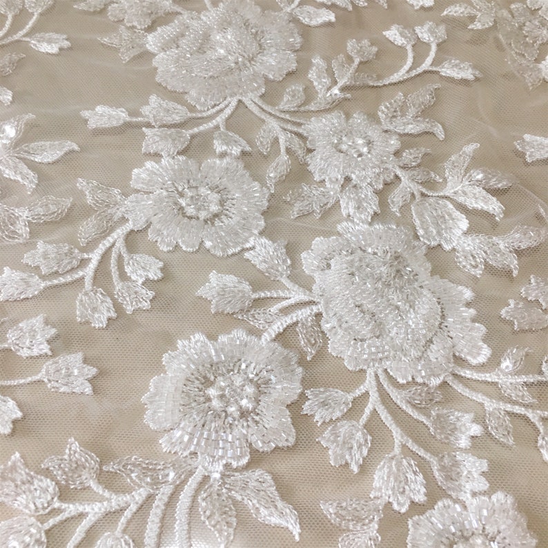 May include: White beaded floral lace fabric with a delicate pattern. The lace is made of small white beads that are sewn together to create a floral design. The fabric is sheer and has a soft, flowing texture.