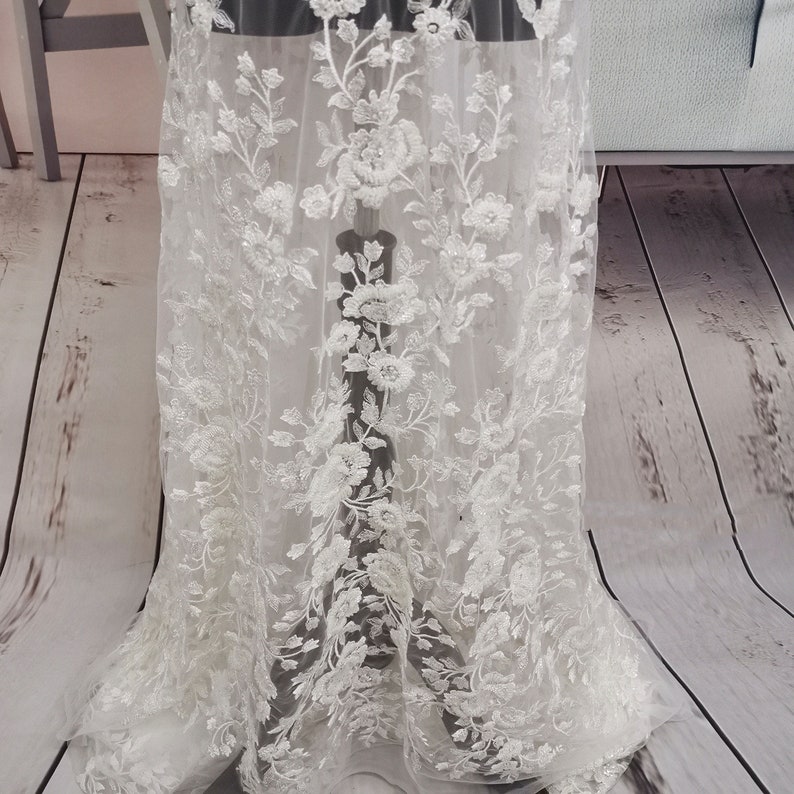 May include: White floral lace fabric with delicate beading and embroidery. The lace features a repeating pattern of flowers and leaves.