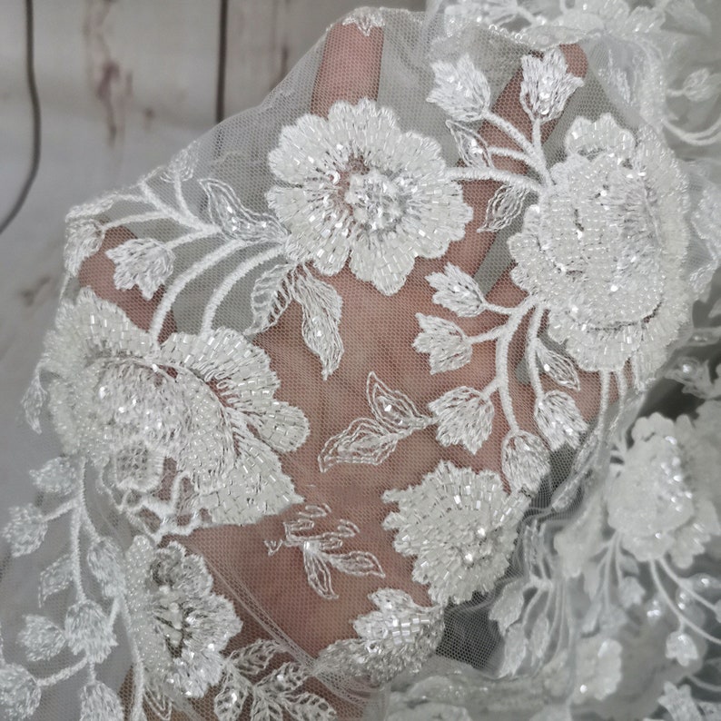 May include: White beaded floral lace fabric with a delicate pattern. The lace is sheer and has a soft, flowing texture.