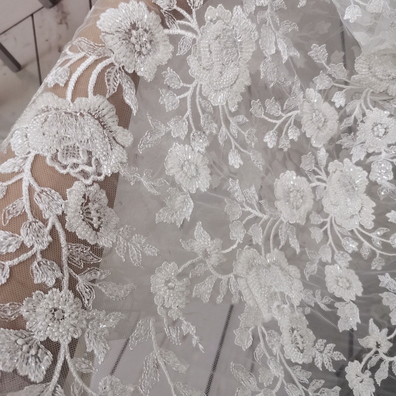 May include: White beaded floral lace fabric with a delicate pattern. The lace is sheer and has a soft, flowing texture.