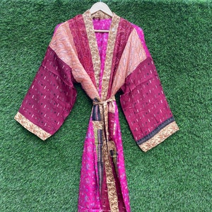 Puede incluir: Una larga bata de seda rosa, roja y dorada con un estampado floral. La bata tiene un cinturón de lazo y mangas largas.