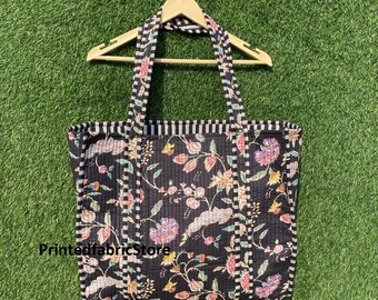 Op de afbeelding: Zwarte quilted tote bag met bloemenprint en wit-zwarte gestreepte afwerking. De tas heeft twee lange hengsels en een platte bodem.