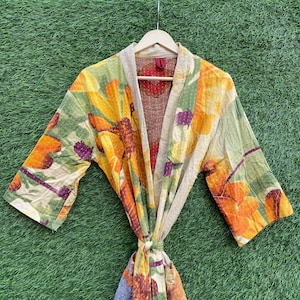May include: A floral print kimono robe with a tie closure. The robe features a vibrant pattern of orange, yellow, and green flowers on a light background. The robe is hanging on a wooden hanger against a green background.