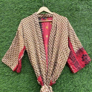 May include: A patterned kimono robe with a beige background and brown elephant motifs. The robe has red accents on the sleeves and edges. The interior is a vibrant red with yellow floral patterns. The robe is hanging on a wooden hanger.