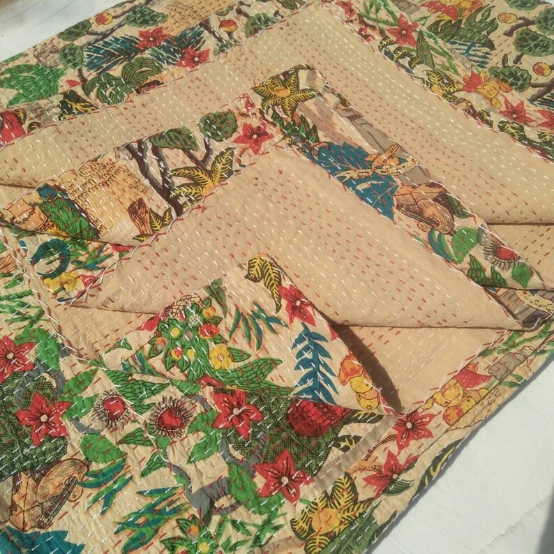 Puede incluir: Una colcha kantha beige con estampado floral y de p&aacute;jaros. La colcha est&aacute; doblada en tres partes, mostrando el intrincado detalle de la costura a mano.
