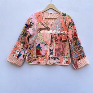 Op de afbeelding: Een roze gewatteerd jack met een bloemenprint en een knoopsluiting. Het jack heeft een patchwork-ontwerp en een wit en roze gestreepte afwerking.