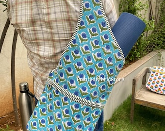 Puede incluir: Una esterilla de yoga azul se lleva en una bolsa de yoga turquesa con un estampado floral. La bolsa tiene una correa y un ribete a rayas blancas y negras. Una botella de agua de acero inoxidable es visible en el fondo.