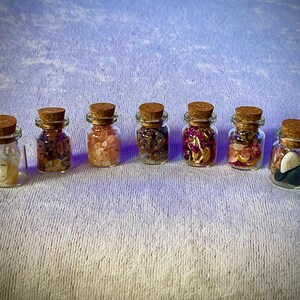 Pode incluir: Sete pequenos frascos de vidro com rolhas de cortiça, cada um preenchido com pedras e cristais de cores diferentes. Os frascos estão dispostos em fila, mostrando uma variedade de elementos naturais. Os conteúdos variam de transparente a rosa e castanho.
