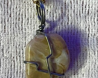 May include: A pendant necklace featuring a polished, light brown stone wrapped in silver-colored wire. The stone has a marbled appearance. The necklace chain is gold-colored. The jewelry is displayed against a white, textured background.