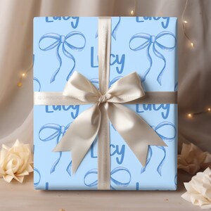 May include: Light blue gift wrap with a white ribbon and a repeating pattern of blue bows and the name "Lucy" in white.
